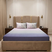 A plush bed with a gradient navy blue and white King Size Percale Ombre 250 TC Bedsheet + Pillow Cover Pair Set, flanked by two nightstands—one with a clock and the other with flowers.