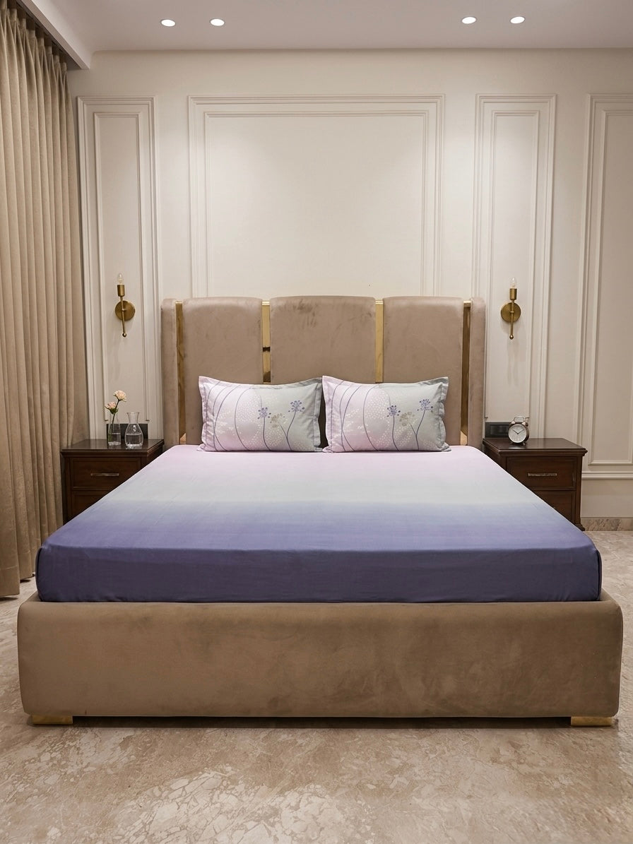 A plush bed with a gradient navy blue and white King Size Percale Ombre 250 TC Bedsheet + Pillow Cover Pair Set, flanked by two nightstands—one with a clock and the other with flowers.